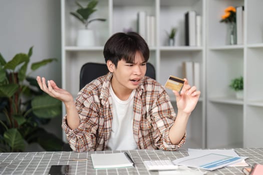 A young man looks frustrated while holding a credit card, reflecting confusion over his finances in a modern home office.