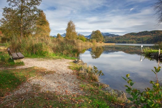 Wide-angle landscape view over the lake Weissensee with wooden bench on the left bank side and the first foothills of the high Alps in the background, near the city Fuessen.