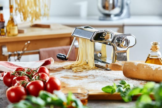 Metallic noodle maker sitting on board in front of kitchen with rolling pin and vegetables in foreground
