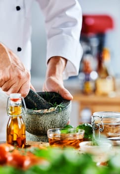 Adult male chef using mortar and pestle to grind random herbs behind blurry foodstuff in the foreground