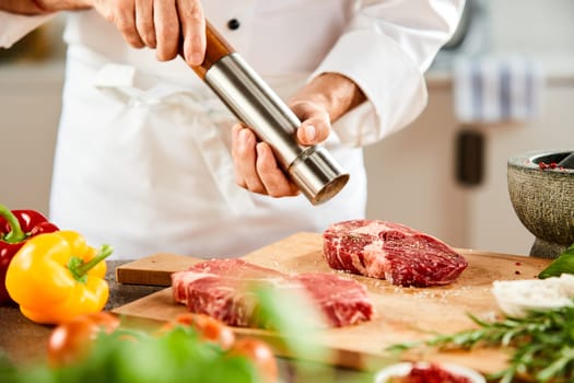 Side view of male chef in white jacket grinding pepper for salted meat net to vegetables in the foreground