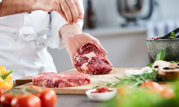 Chef sprinkling cooking salt onto a raw ribeye steak while preparing dinner with fresh salad ingredients and herbs on the counter