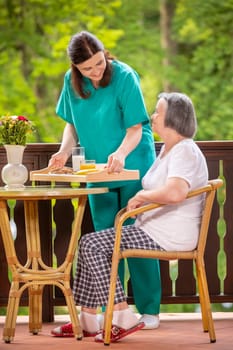 Happy nurse or caregiver serving healthy breakfast to elderly woman on the terrace at nursing home.
