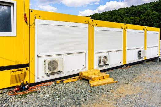 White air conditioning unit on the back side of bright yellow colored building with large green forest and blue sky in background