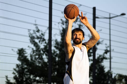 The smiling playing basketball player is throwing the basketball ball outdoor.