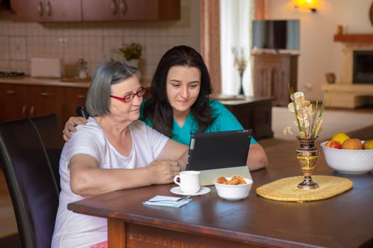 Female nurse and senior woman using tablet PC in kitchen at nursing home.
