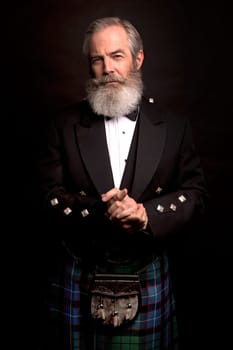 close up of senior man with grey hair and full beard, wearing scotting kilt on dark background