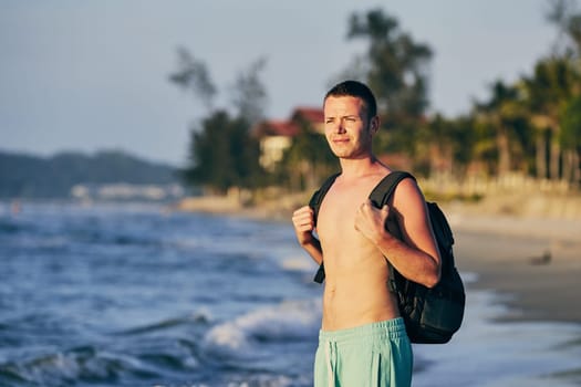 Young man (tourist) with backpack standing on sand beach.