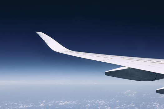 Wing of modern airplane flying over clouds against dark blue sky.