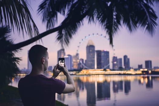 Young man (tourist) taking photo of the city by mobile phone. Urban skyline of Singapore at sunset.