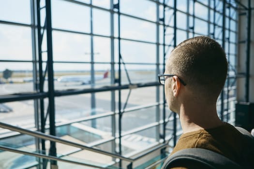 Young passenger waiting at airport and looking through window to airplane.