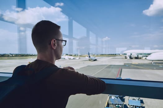 Young passenger waiting at airport and looking through window to airplane.