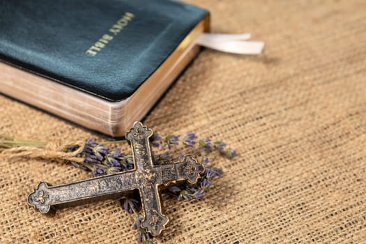 Closeup of metalic Christian cross on lavanda flowers next to Holy Bible on textile background.