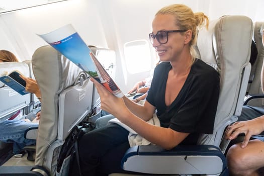 Cheerful woman reading magazine on airplane during flight. Female traveler reading seated in passanger cabin.