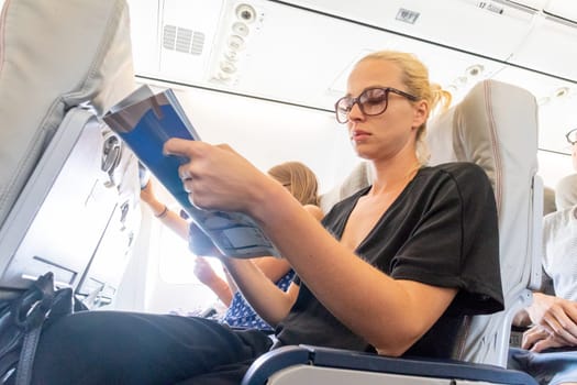 Cheerful woman reading magazine on airplane during flight. Female traveler reading seated in passanger cabin.