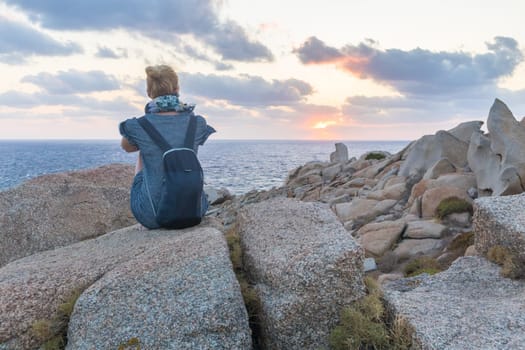 Solitary young female traveler watches a beautiful sunset on spectacular rocks of Capo Testa, Sardinia, Italy, a popular summer traveling destination in Europe.