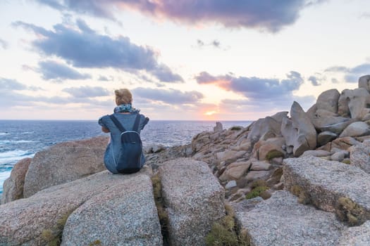 Solitary young female traveler watches a beautiful sunset on spectacular rocks of Capo Testa, Sardinia, Italy, a popular summer traveling destination in Europe.
