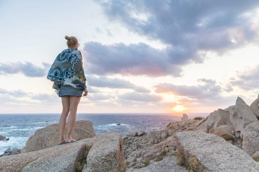Solitary young female traveler watches a beautiful sunset on spectacular rocks of Capo Testa, Sardinia, Italy, a popular summer traveling destination in Europe.