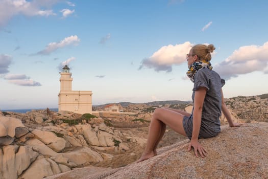 Solitary young female traveler watches a beautiful sunset over lighthouse and spectacular rock formations of Capo Testa, Sardinia, Italy, a popular summer traveling destination in Europe.