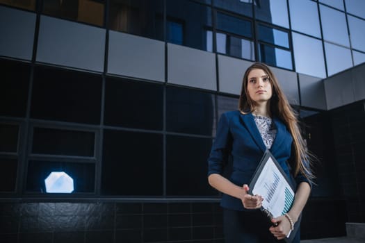 Successful beautiful young business woman standing against a background of buildings and holding folder.