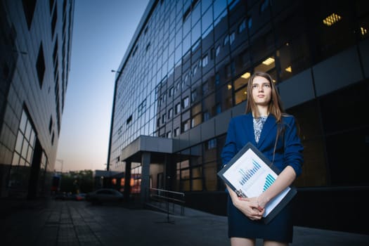 Successful beautiful young business woman standing against a background of buildings and holding folder.
