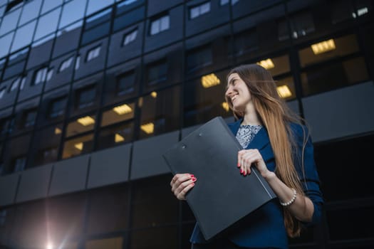 Successful beautiful young business woman standing against a background of buildings and holding folder.