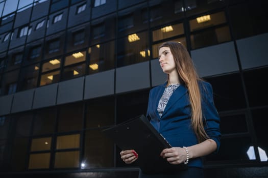 Successful beautiful young business woman standing against a background of buildings and holding folder.