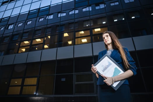 Successful beautiful young business woman standing against a background of buildings and holding folder.