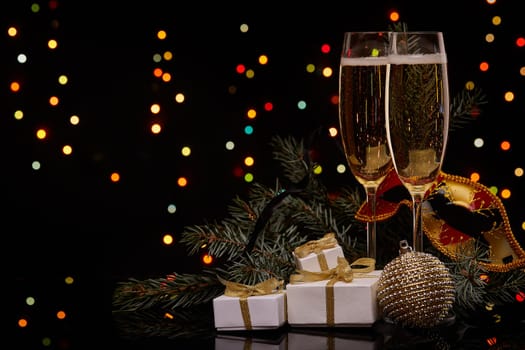 Two glasses with champange, fir tree branch with decoration, gift boxes on a black background with multi-colored lightes of garland. New year and Christmas.