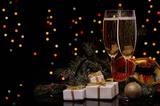 Two glasses with champange, fir tree branch with decoration, gift boxes on a black background with multi-colored lightes of garland. New year and Christmas.