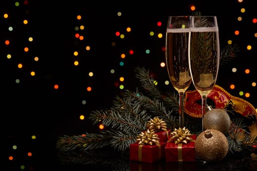 Two glasses with champange, fir tree branch with decoration, gift boxes on a black background with multi-colored lightes of garland. New year and Christmas.