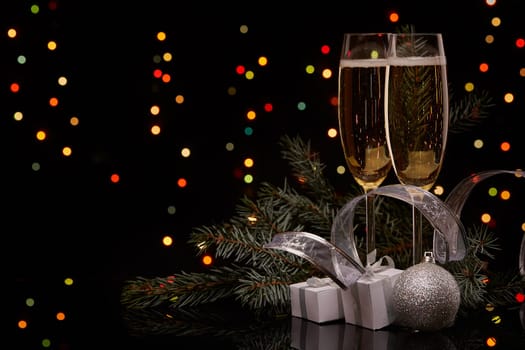 Two glasses with champange, fir tree branch with decoration, gift boxes on a black background with multi-colored lightes of garland. New year and Christmas.