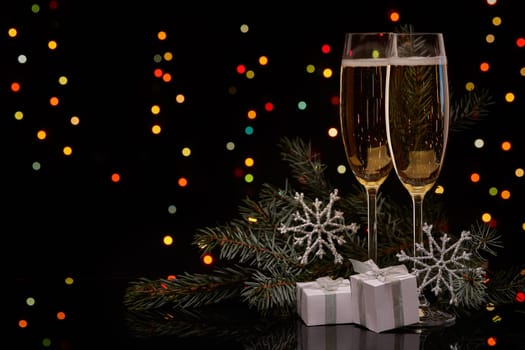 Two glasses with champange, fir tree branch with decoration, gift boxes on a black background with multi-colored lightes of garland. New year and Christmas.