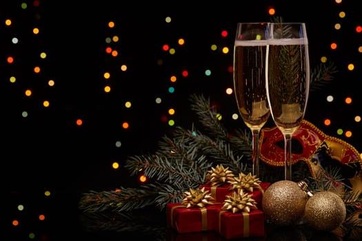 Two glasses with champange, fir tree branch with decoration, gift boxes on a black background with multi-colored lightes of garland. New year and Christmas.