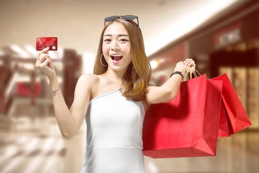 Cheerful asian woman with red shopping bags showing her credit card on the shopping mall