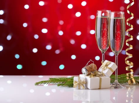 Two glasses with champange, fir tree branch, gift boxes and christmas decorations on a red background with lights of garland. New year and Christmas.