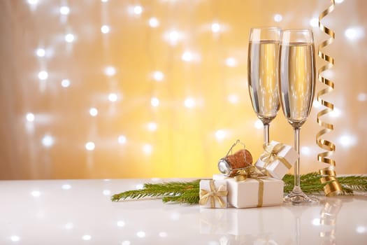 Two glasses with champange, fir tree branch, gift boxes and christmas decorations on a yellow background with lights of garland. New year and Christmas.