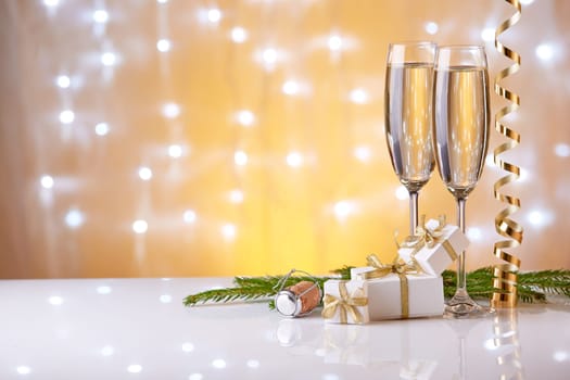 Two glasses with champange, fir tree branch, gift boxes and christmas decorations on a yellow background with lights of garland. New year and Christmas.
