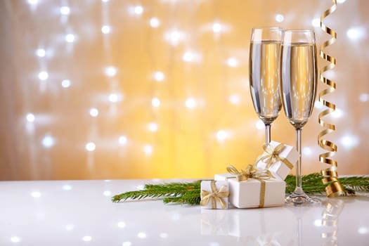 Two glasses with champange, fir tree branch, gift boxes and christmas decorations on a yellow background with lights of garland. New year and Christmas.