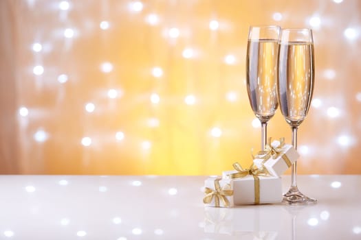 Two glasses with champange and gift boxes on a yellow background with lights of garland.