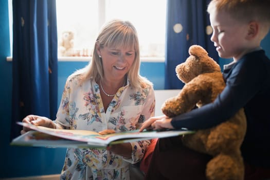 Grandmother and her grandson choosing a book to read before bedtime. The little boy is holding his teddy bear and pointing in the book.