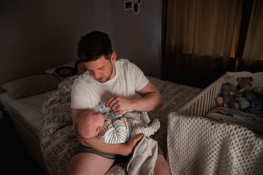 Father checking on his newborn baby boy at night as his wife stays asleep in bed.