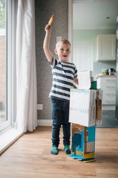 Little boy standing indoors next to a home made robot he has made out of old cardboard boxes. He is holding a rocket ice lolly and pretedning that it is flying.