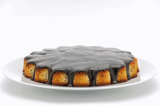 Round cake with poppy seeds covered in chocolate on a plate on a white background.