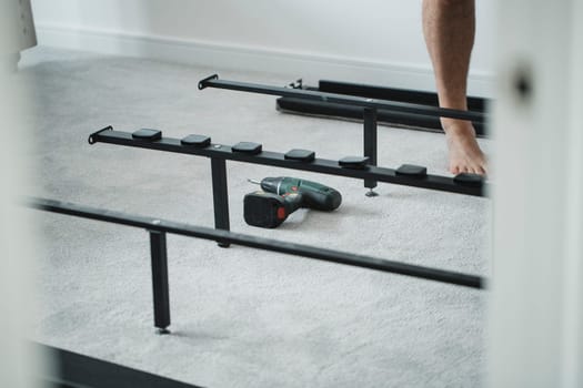 A powerdrill is lying on the carpet near a bed frame which a man is putting together in a new home.