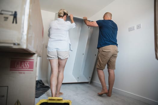 Mature couple are putting a wardrobe up in a bedroom of their new home.