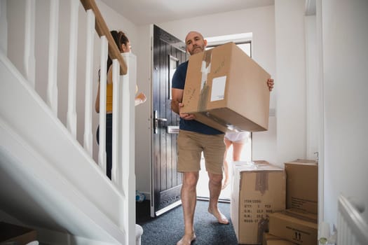 Mature man is carrying boxes in to his new home. His daughter and wife can be seen behind him.