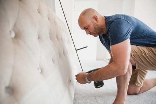 Mature man is building a bed in his new home with a power drill.