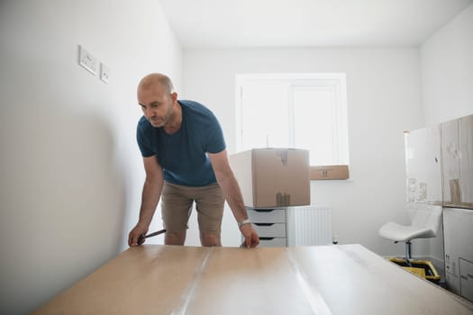 Mature man is moving furniture in to a new home. He is building a wardrobe in one of the bedrooms.
