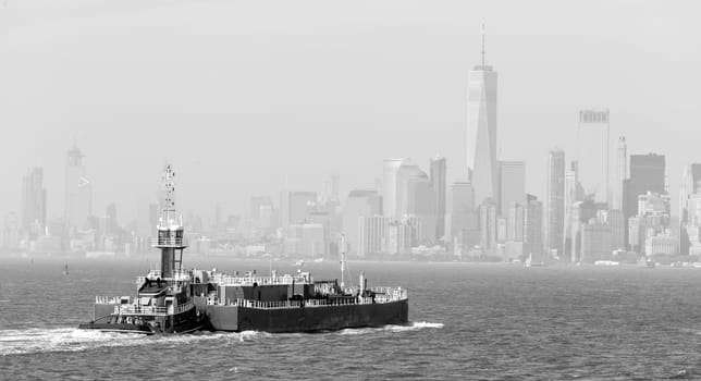 Freight tug pushing cargo ship to the port in New York City and Lower Manhattan skyscarpers skyline in background. New York City, USA. Black and white image.